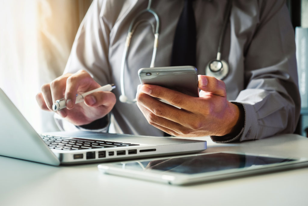 doctor working with mobile phone and stethoscope and digital tablet laptop in modern office at hospital in morning light